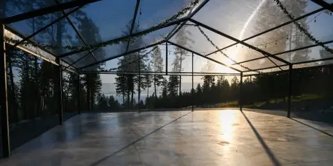 Open-sided glass wedding pavilion with concrete floor and string lights overlooking a lake and forest at sunset