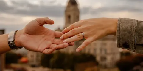 A close-up of two hands exchanging a diamond ring with the Old Courthouse tower in the background.