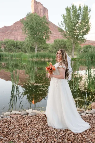 A bride in a white wedding dress and veil stands on a rocky shore holding a bouquet of orange flowers, with a pond and red rock cliffs in the background.