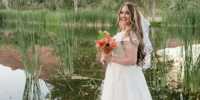 A bride in a white wedding dress and veil stands on a rocky shore holding a bouquet of orange flowers, with a pond and red rock cliffs in the background.