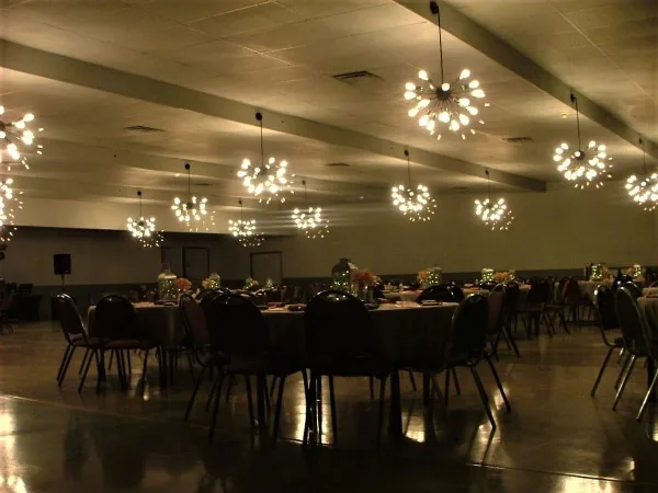 Interior view of a large event hall with round tables, chairs, and hanging light fixtures.