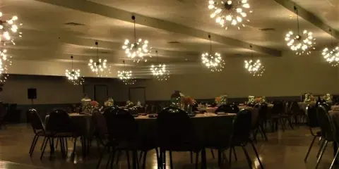 Interior view of a large event hall with round tables, chairs, and hanging light fixtures.