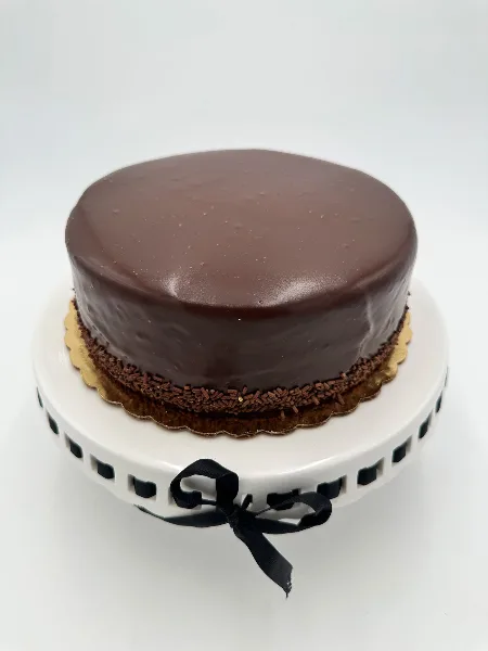 A round chocolate cake with a smooth glaze and chocolate sprinkles at the base, presented on a white cake stand.