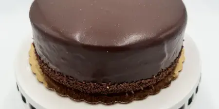 A round chocolate cake with a smooth glaze and chocolate sprinkles at the base, presented on a white cake stand.