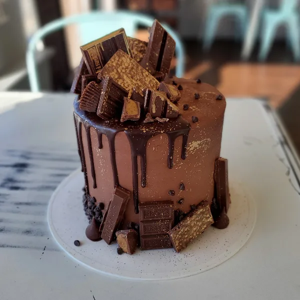A chocolate drip cake decorated with various chocolate bars and toppings on a white cake stand.