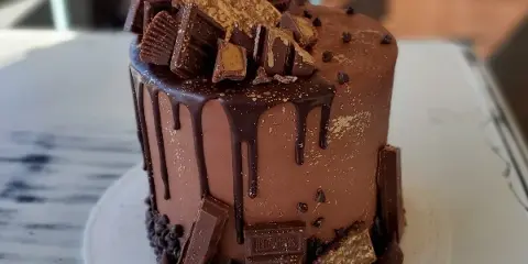 A chocolate drip cake decorated with various chocolate bars and toppings on a white cake stand.