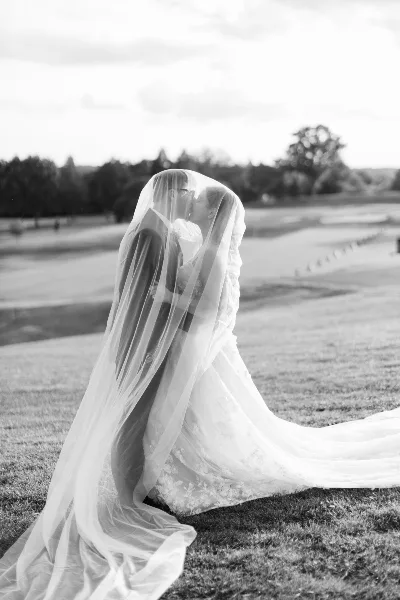 Black and white photo of a bride and groom kissing under a veil on a grassy field.