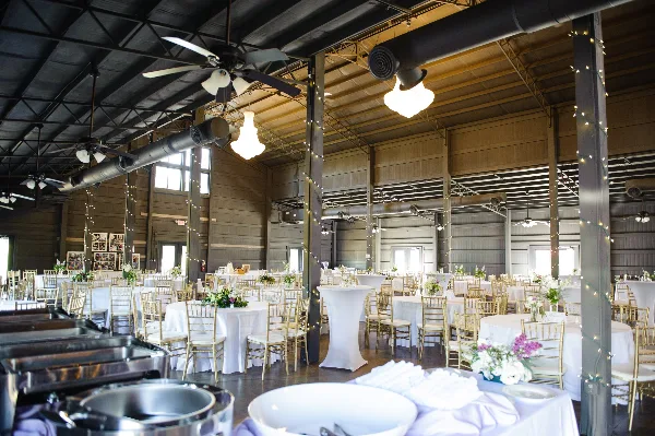 A spacious industrial-style wedding venue interior featuring round tables with white linens, gold chiavari chairs, and buffet stations.