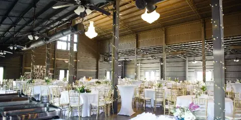 A spacious industrial-style wedding venue interior featuring round tables with white linens, gold chiavari chairs, and buffet stations.