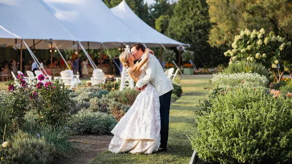 A bride and groom kissing in a garden with a white wedding tent and floral arrangements in the background.