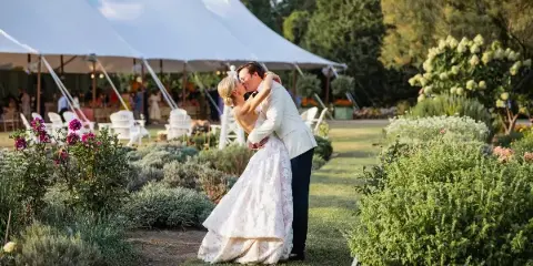 A bride and groom kissing in a garden with a white wedding tent and floral arrangements in the background.
