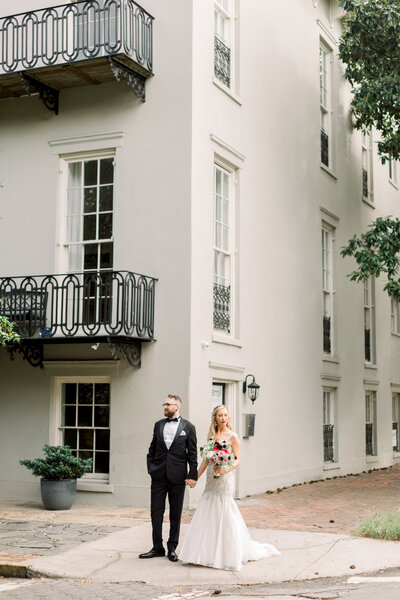 A bride and groom standing together on a sidewalk in front of a historic building with balconies.