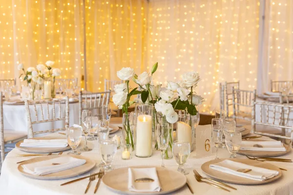 Elegant wedding reception table setting with white roses, candles, and gold cutlery against a backdrop of string lights.