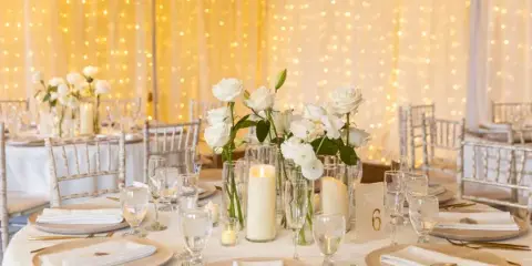 Elegant wedding reception table setting with white roses, candles, and gold cutlery against a backdrop of string lights.