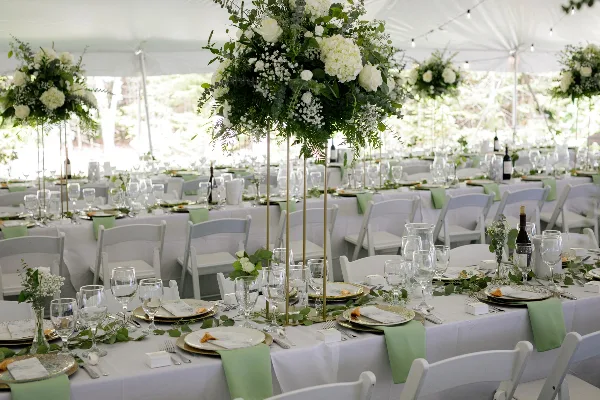 A wedding reception setup with white tables, chairs, and tall floral centerpieces under a white tent.