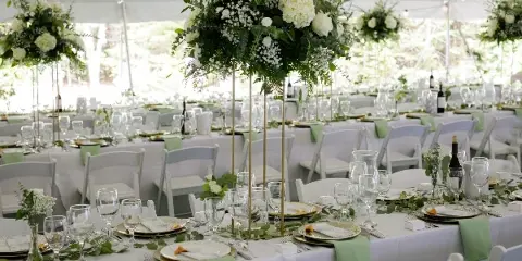 A wedding reception setup with white tables, chairs, and tall floral centerpieces under a white tent.