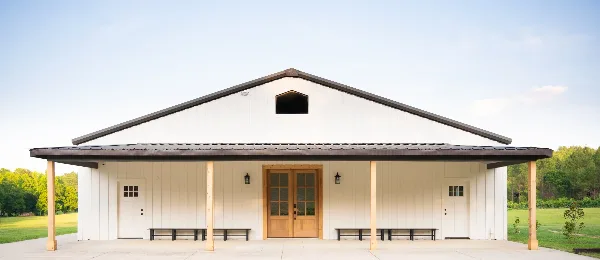 Exterior view of a modern white building with a dark roof and wooden porch, set against a green field and trees.