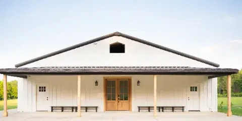 Exterior view of a modern white building with a dark roof and wooden porch, set against a green field and trees.