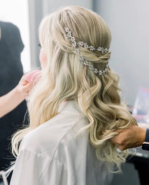 A close-up view of a hairstylist styling a client's blonde hair into an elegant half-up, half-down wedding hairstyle with a jeweled hair accessory.