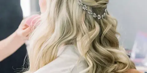 A close-up view of a hairstylist styling a client's blonde hair into an elegant half-up, half-down wedding hairstyle with a jeweled hair accessory.