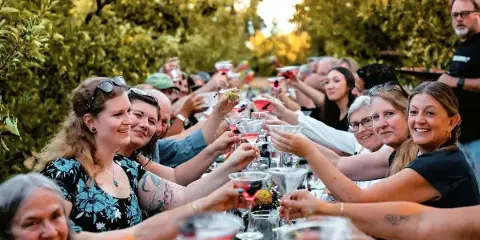 A group of smiling guests at an outdoor event raising cocktail glasses in a toast.