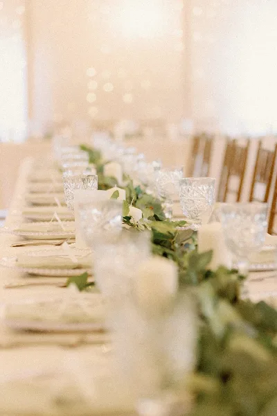 Indoor wedding banquet table set with white linens, crystal glassware, candles, and a greenery runner in front of chiavari chairs.