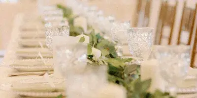Indoor wedding banquet table set with white linens, crystal glassware, candles, and a greenery runner in front of chiavari chairs.