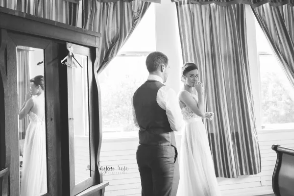 A black and white documentary-style photograph of a bride and groom sharing an emotional moment in a room with striped curtains, featuring the Bayou Rose Photography watermark.