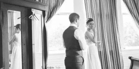 A black and white documentary-style photograph of a bride and groom sharing an emotional moment in a room with striped curtains, featuring the Bayou Rose Photography watermark.