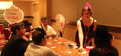 A performer in a black dress and hat interacts with guests seated at a table during an indoor event.