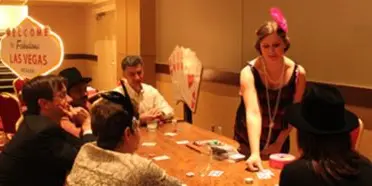A performer in a black dress and hat interacts with guests seated at a table during an indoor event.