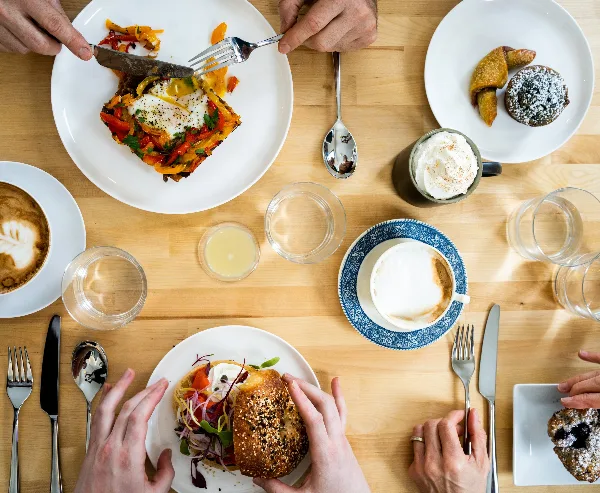 A top-down view of a wooden table set with plates of savory dishes, pastries, and cups of coffee, with several people eating together.