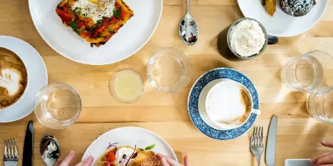 A top-down view of a wooden table set with plates of savory dishes, pastries, and cups of coffee, with several people eating together.