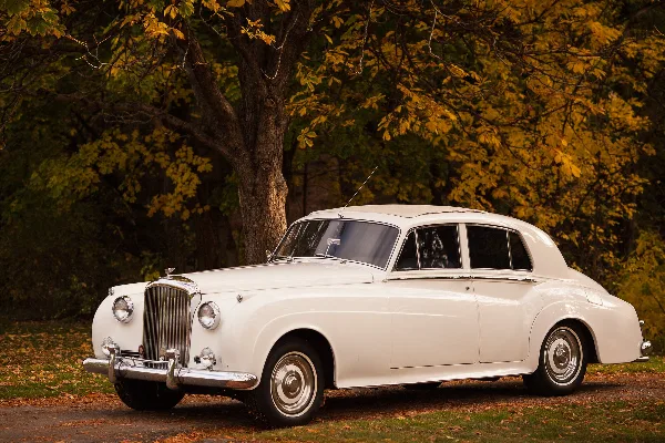 A white vintage luxury sedan parked on a gravel path with autumn trees in the background.