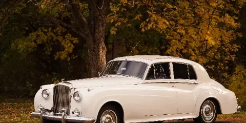A white vintage luxury sedan parked on a gravel path with autumn trees in the background.