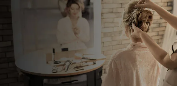 A bridal hair stylist styling an updo with pearl accessories on a bride in a robe, with makeup tools on a table.