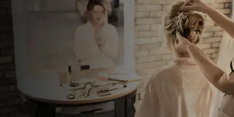 A bridal hair stylist styling an updo with pearl accessories on a bride in a robe, with makeup tools on a table.