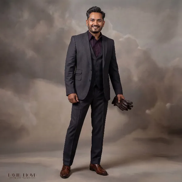 A professional studio portrait of a man in a dark suit and brown shoes, holding leather gloves, standing against a textured grey background.