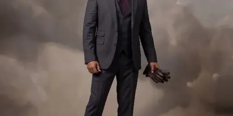 A professional studio portrait of a man in a dark suit and brown shoes, holding leather gloves, standing against a textured grey background.