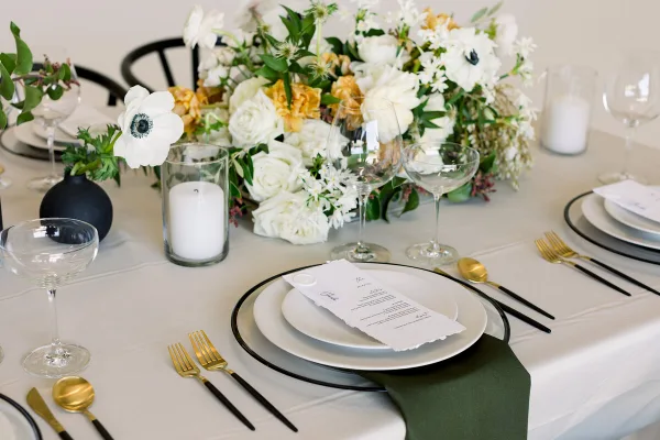 Elegant wedding table setting with white and yellow floral centerpiece, gold and black flatware, and place cards on a white tablecloth.