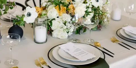 Elegant wedding table setting with white and yellow floral centerpiece, gold and black flatware, and place cards on a white tablecloth.