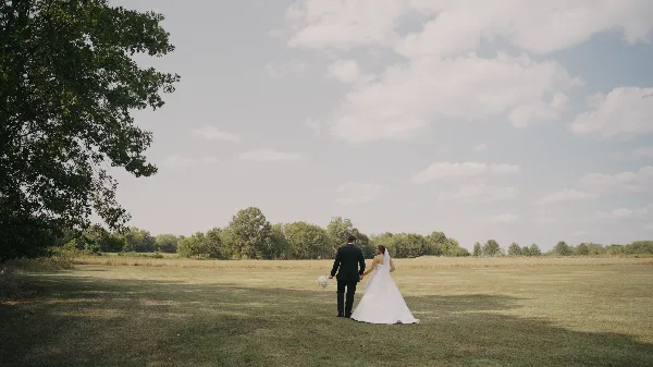 A bride and groom walking hand-in-hand across a grassy field under a cloudy sky.