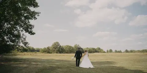 A bride and groom walking hand-in-hand across a grassy field under a cloudy sky.