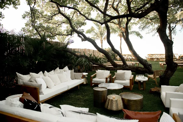 Outdoor event lounge with white cushioned sofas, armchairs, and metallic coffee tables arranged on grass under a large tree.