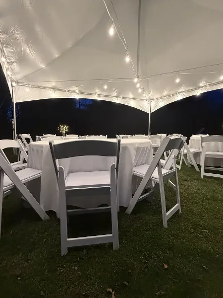 A white event tent set up on a lawn at night, featuring round tables with white linens and white folding chairs.