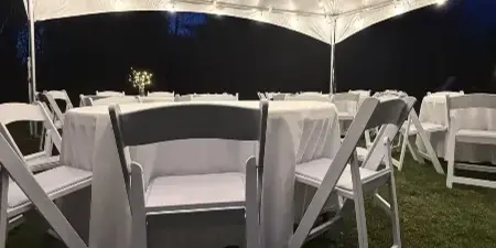 A white event tent set up on a lawn at night, featuring round tables with white linens and white folding chairs.