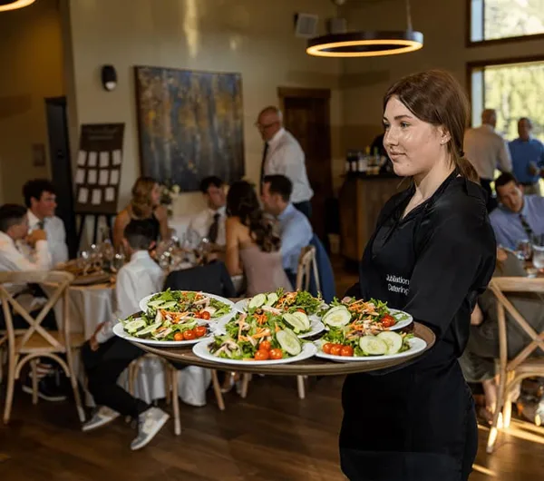 A server in a Jubilations Catering apron carries a tray of plated salads to a dining room full of guests.