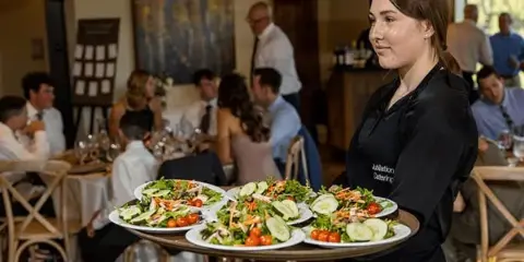 A server in a Jubilations Catering apron carries a tray of plated salads to a dining room full of guests.
