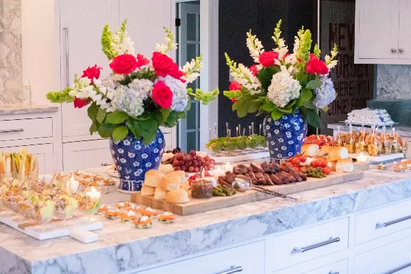 A catering buffet table set with a charcuterie board, sliced meats, bread rolls, and large floral centerpieces in blue vases.