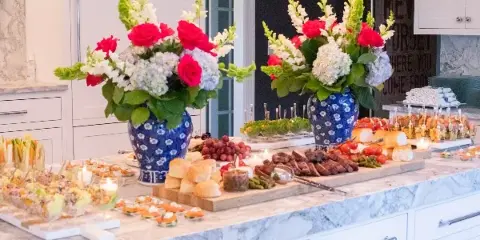 A catering buffet table set with a charcuterie board, sliced meats, bread rolls, and large floral centerpieces in blue vases.